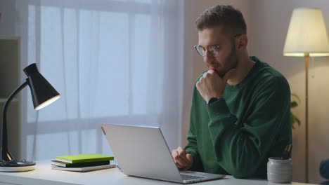 Un-Joven-Con-Gafas-Envía-Un-Mensaje-En-Un-Chat-En-Línea-Y-Espera-Una-Respuesta-Usando-Internet-Por-La-Noche-En-Casa.-Un-Retrato-Masculino-Con-Un-Cuaderno.